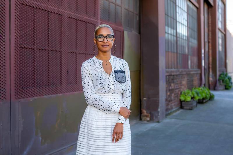 Woman in a white dress and glasses poses for a photo outside of a warehouse. 