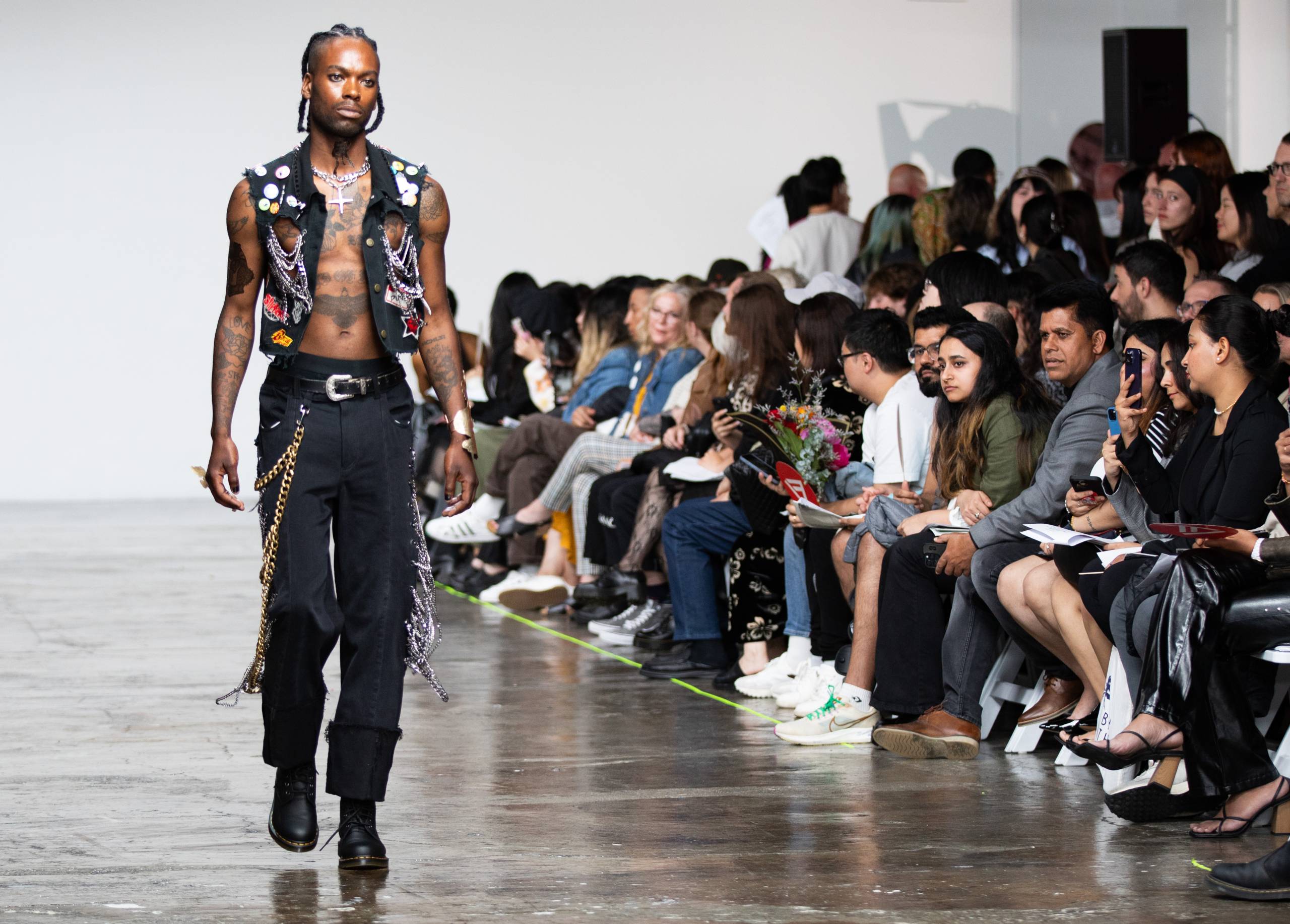 Black man in vest and black pants with chains and rivets walks in front of seated crowd