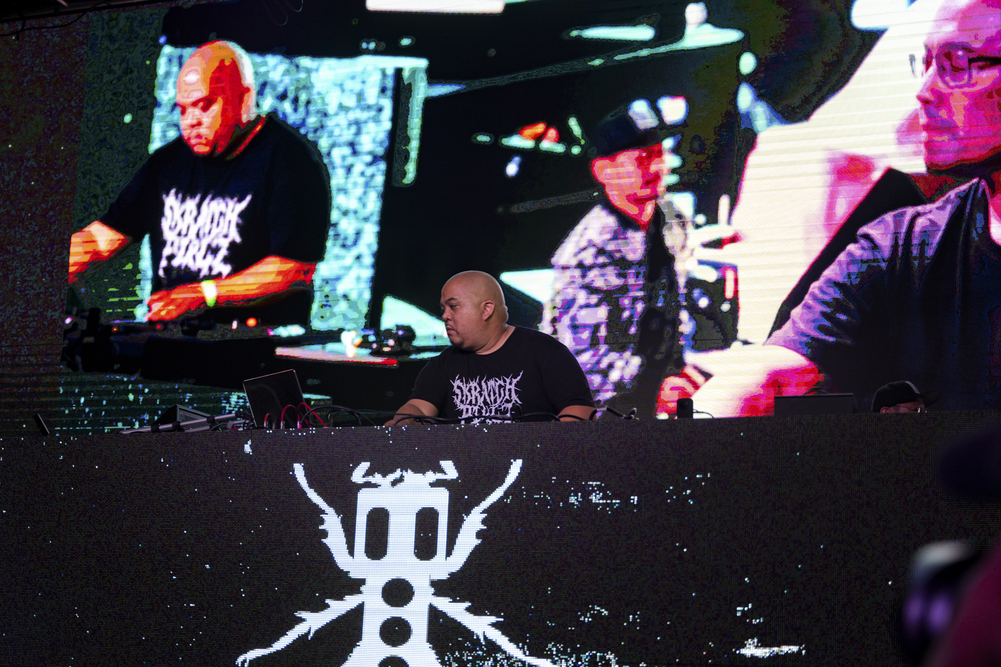 A person with a shaved head stands at a table as a screen behind them shows the images of several people.