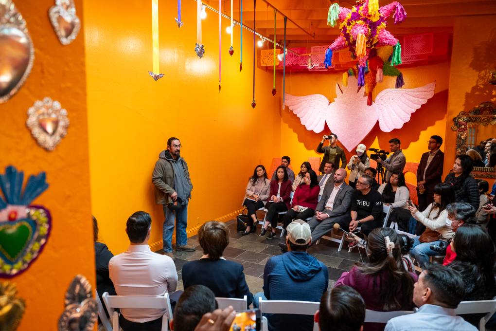 A person with a goatee stands in front of a large group of people sitting in fold-out chairs in a room painted bright orange.