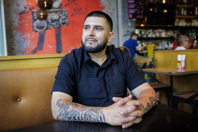 Man with beard and arm tattoos looks off into the distance while seated inside a restaurant.