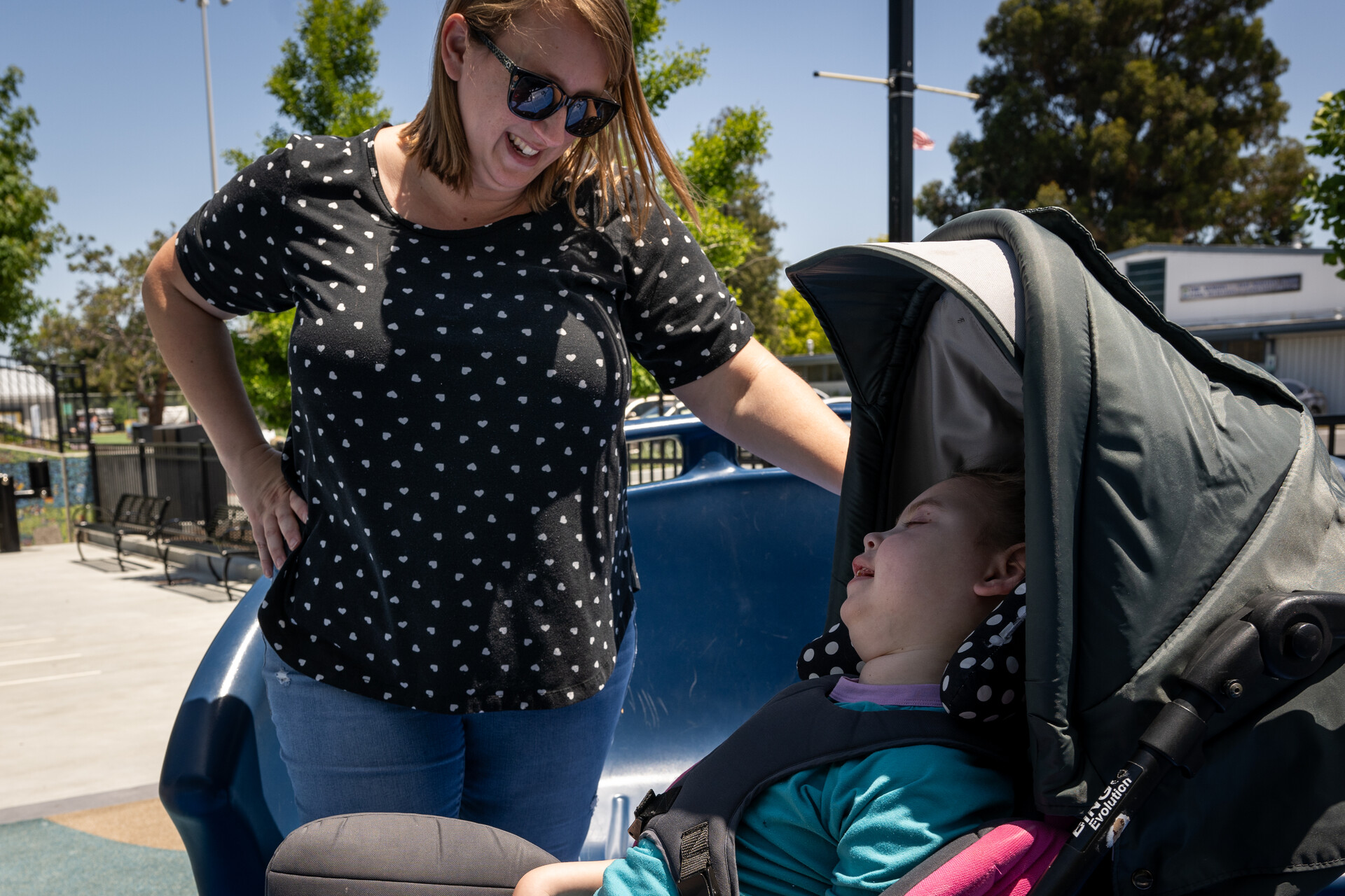 A standing woman smiles down at a smiling child who sits in what appears to be a stroller outdoors in a park