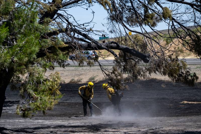 Two people dressed in firefighter equipment use tools to extinguish a fire.