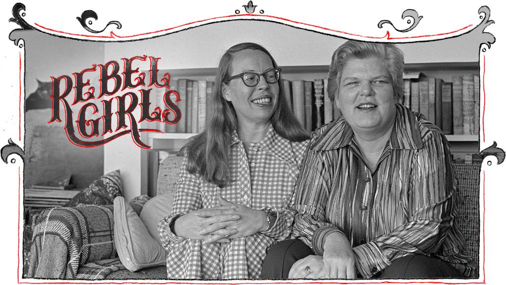 A white bespectacled woman wearing a checked dress smiles warmly at another white woman as they sit close together on a couch. A wall lined with books is behind them.