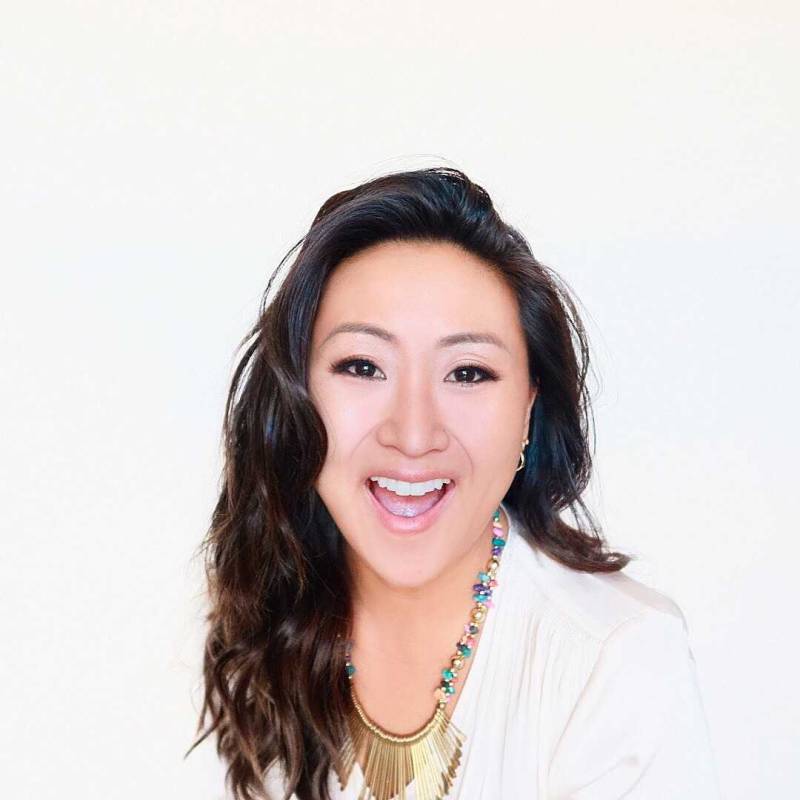 Woman faces camera with a smile, while wearing a gold necklace over a white top in front of a white background. 