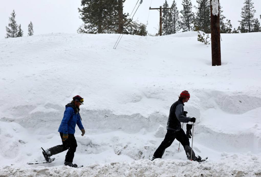 Two people dressed in winter clothing walk next to a large pile of snow, covering trees and electric poles.