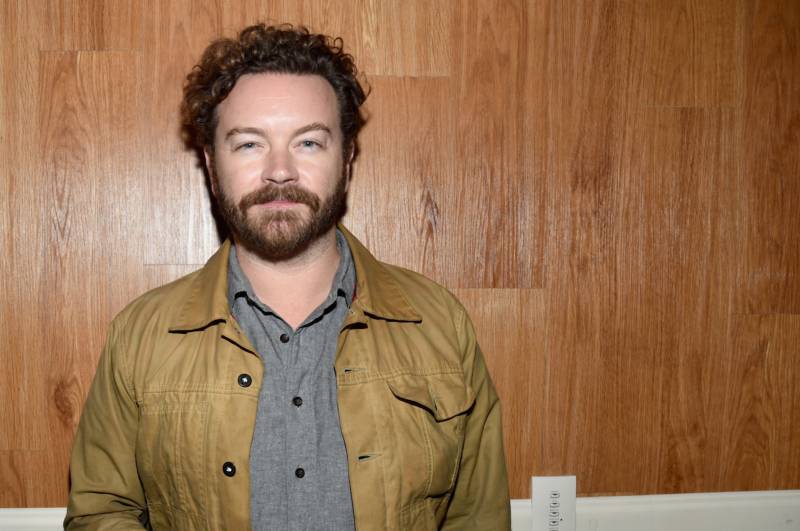 A white man with curly brown hair and beard stands in front a wood-paneled wall. He is wearing a polo shirt and suede jacket.