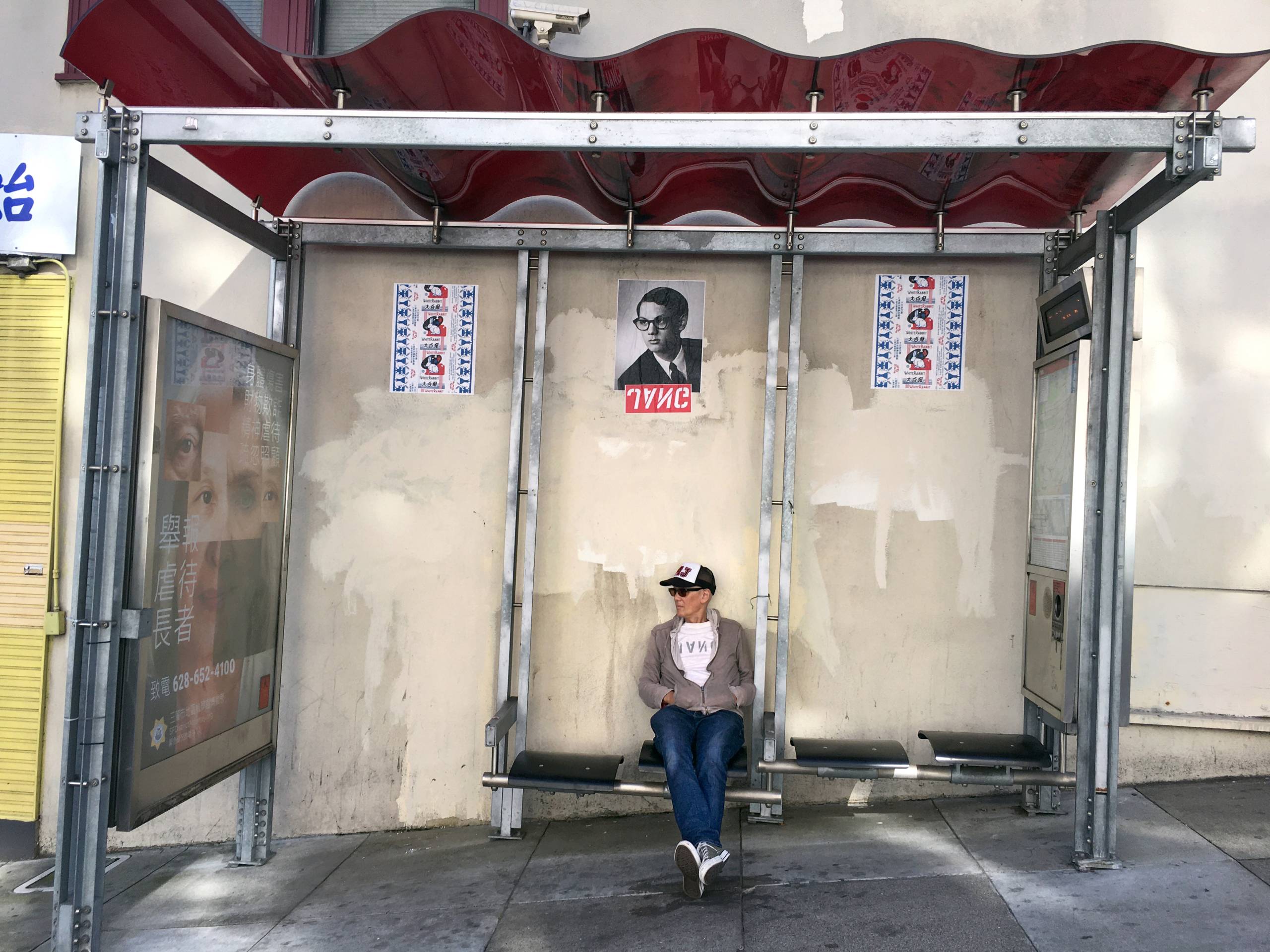 Man sits at bus stop with hands in pockets, wheat paste of "JANG" poster above him on wall